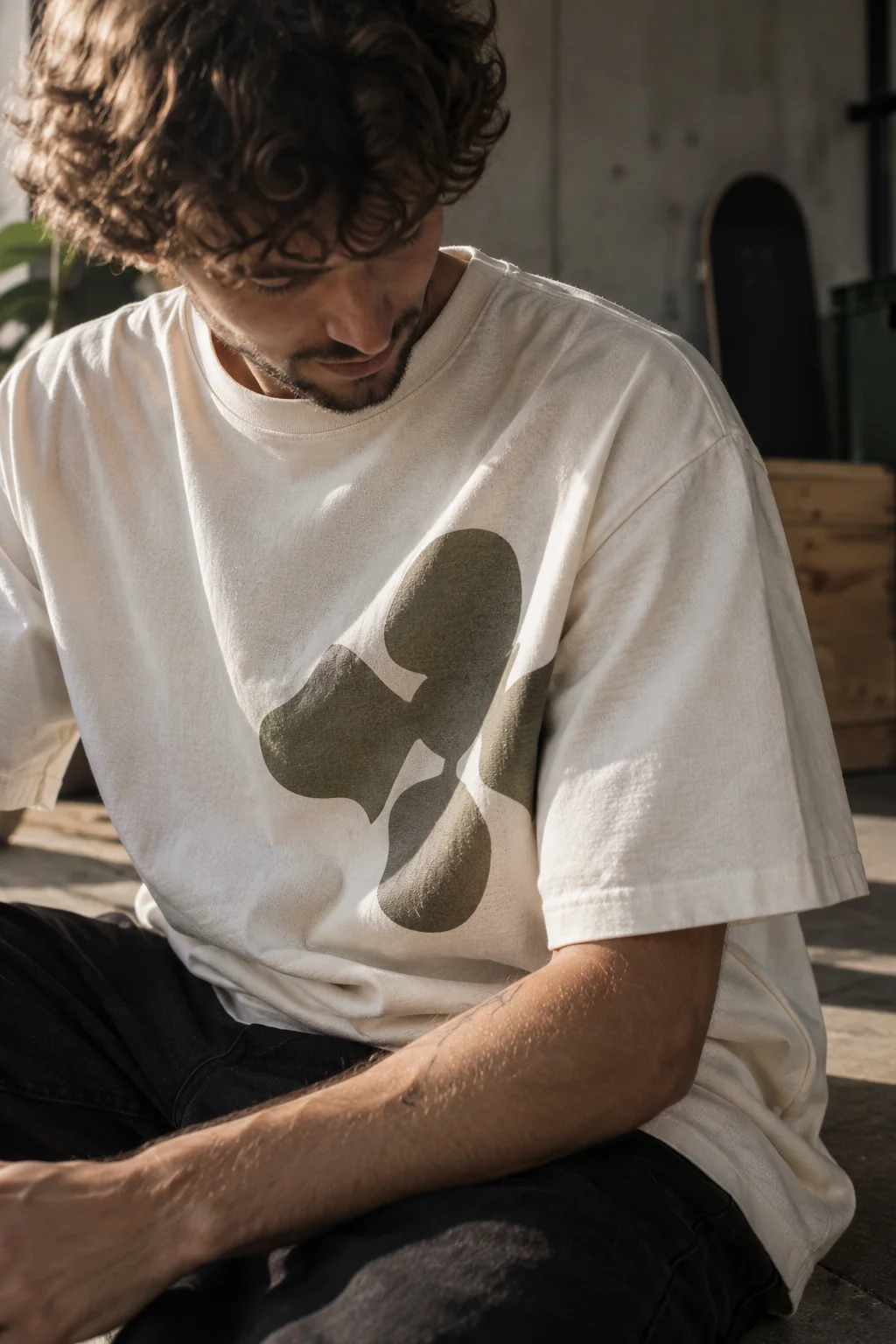 A person in an oversized off-white t-shirt with a large olive abstract print, seated in warm side light